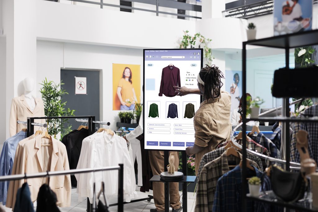 a young man ordering a shirt on a touchscreen monitor in a fashion shop surrounded by many different types and styles of shirt