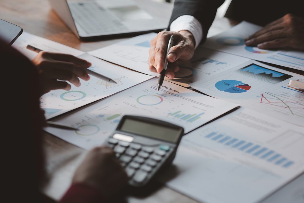 a business group surrounding a monthly budget and expenditure plan, a man is holding a pen to one sheet while a woman holds a digital pen to another, there is a calculator in the foreground with a blank screen