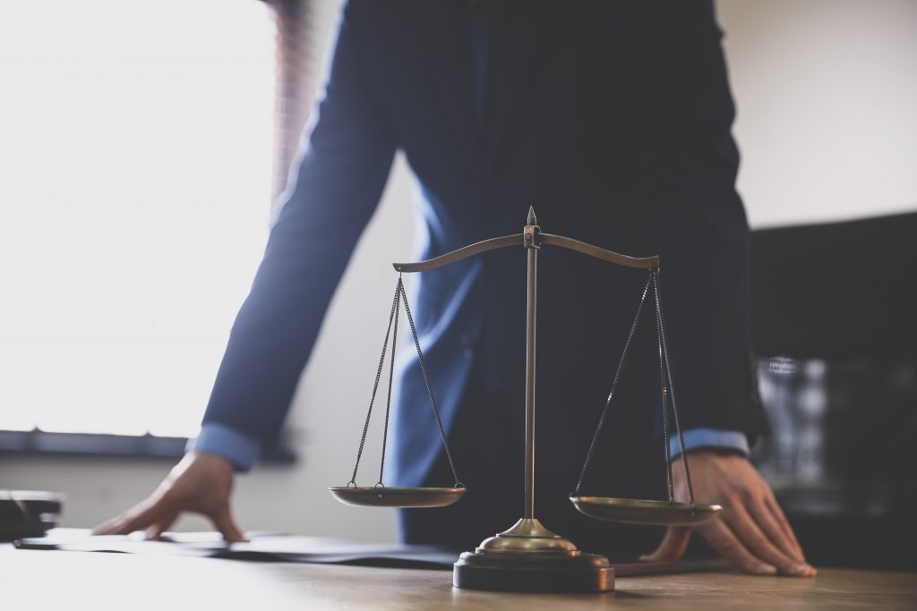 A male lawyer at a table in an office, with a focus on a set of scales of justice. The table is wooden with a black leather chair in the background