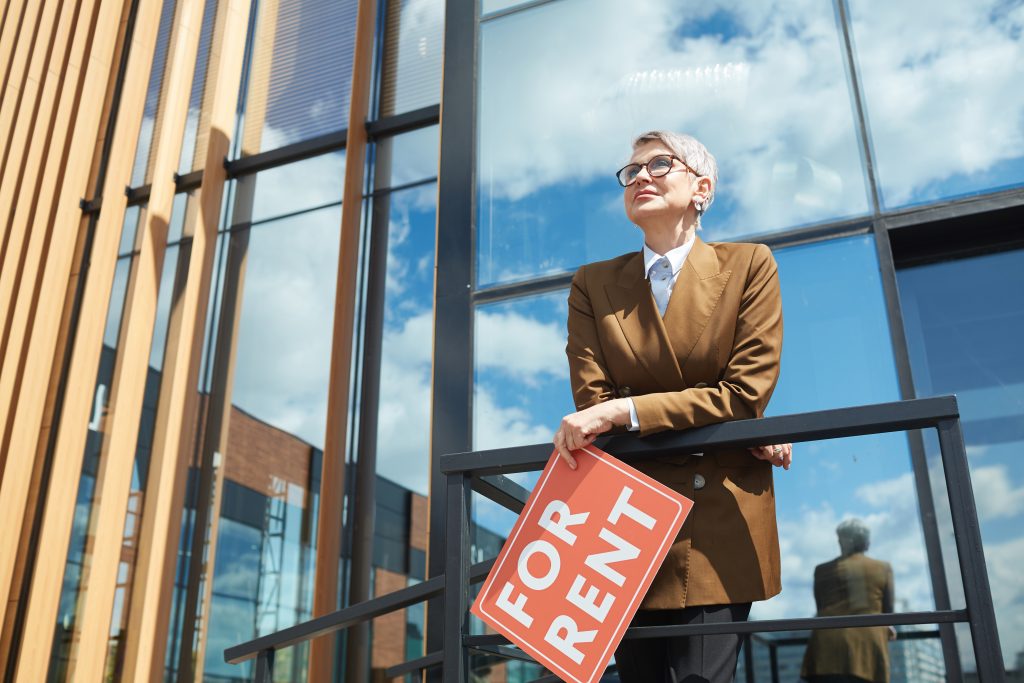 Mature businesswoman in eyeglasses with short, grey hair working as a realtor at modern office building holding a "for rent" sign in a brown business suit