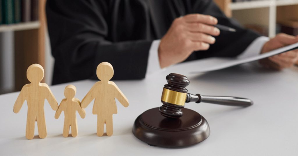 A gavel and hammer next to three wooden figures of a family, with a lawyer writing on a pad in the background